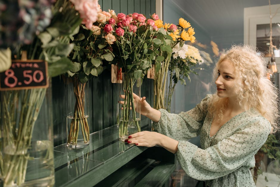 A woman with curly blonde hair and fair skin selecting a floral arrangement in a flower shop. She is wearing a light-colored, long-sleeved blouse with a subtle floral pattern and has red-painted nails