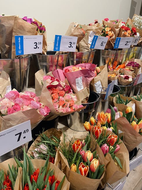 A display of various fresh flower bouquets arranged in metallic buckets and wrapped in brown paper, featuring a colorful mix of roses, tulips, and carnations in shades of pink, orange, red, and purple