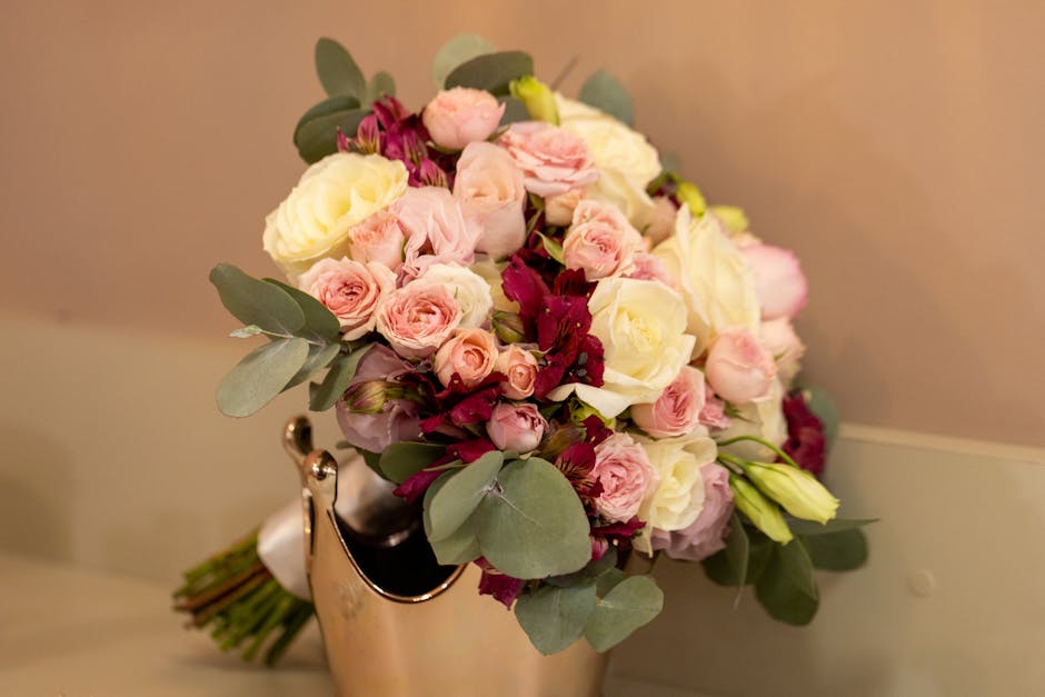 A floral bouquet arrangement featuring a mix of cream and soft pink roses, complemented by deep burgundy flowers and lush green eucalyptus leaves. The bouquet is placed in a metallic gold vase, with t