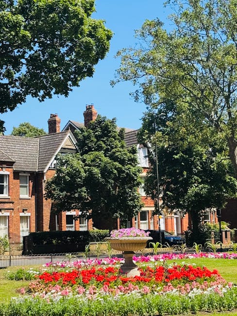 The image features a vibrant flower bed in a park setting, with red, pink, and white blooms arranged in neat, lush patches. Behind the flower bed, there is a large stone planter with a lush green plan