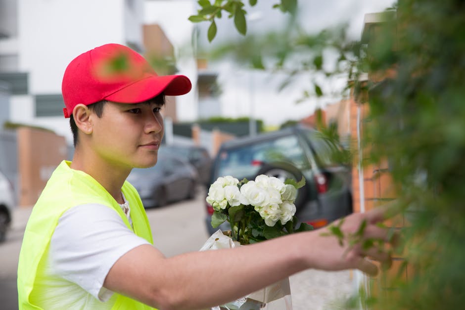 A young delivery person wearing a red cap and a neon yellow vest is standing outdoors on a residential street, holding a bouquet of fresh white hydrangeas wrapped in paper. The arrangement features lu