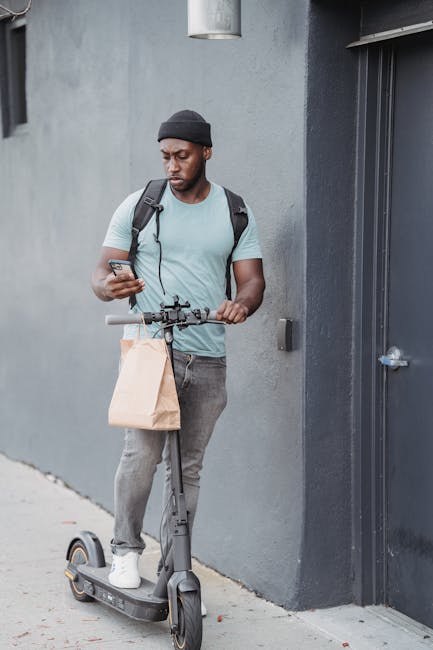 A young man with dark skin, wearing a black beanie, light blue T-shirt, and grey jeans, is standing beside a grey wall next to a metal door. He is holding a smartphone in his right hand, looking at it
